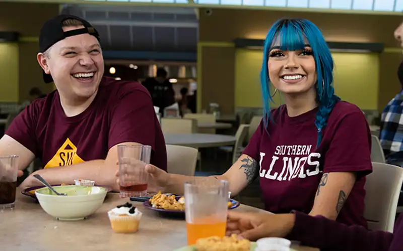 Students sharing a meal in the dining hall, smiling and talking at a table.