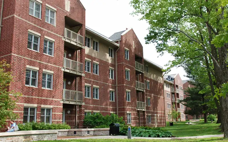 Brick residence hall with multiple floors and small balconies, surrounded by trees and landscaped walkways.