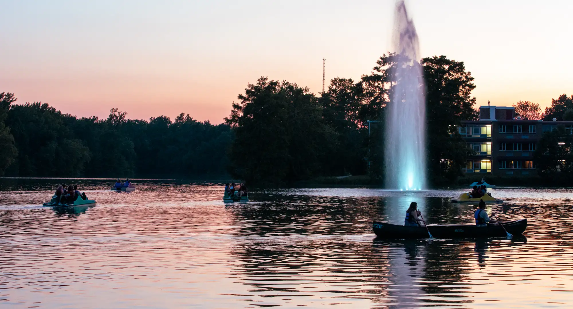 Students canoeing and paddle-boating on the campus lake at sunset with the illuminated fountain and Thompson Point residence halls in the background.