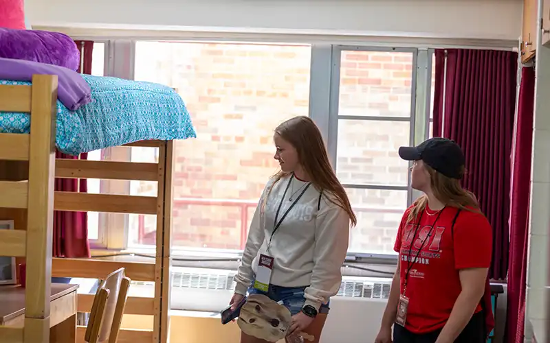 Two students stand inside a residence hall room, looking around as they tour the space with lofted beds, desks, and large windows.