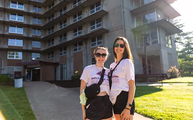Two roommates standing outside an SIU residence hall on a sunny day, smiling and wearing matching Salukis shirts.