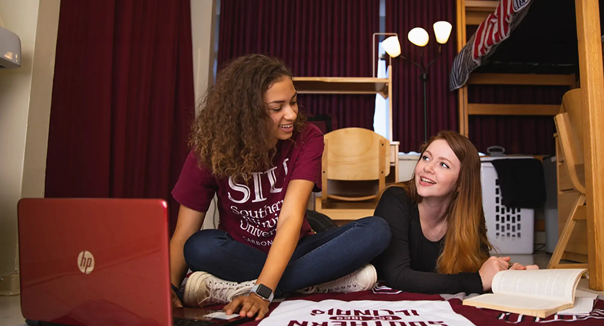 Two students sit on the floor of their residence hall room, smiling and talking while studying with a laptop and an open book nearby.
