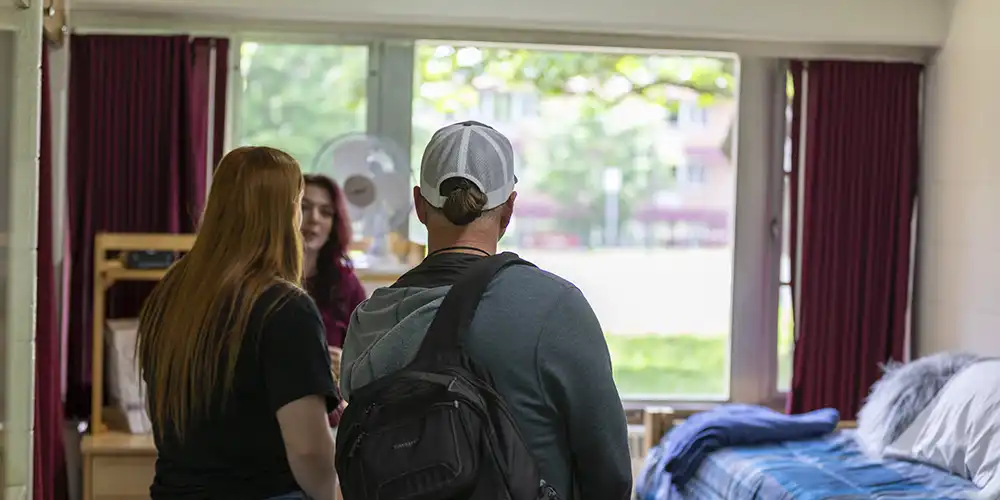 People tour a residence hall room, standing near the window and looking at the setup with beds and shelving.