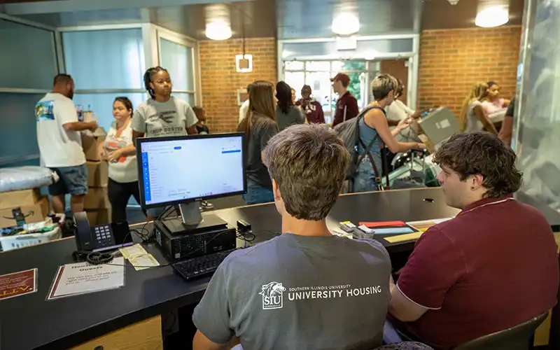 Students check in at a University Housing front desk while others move belongings through the lobby behind them.