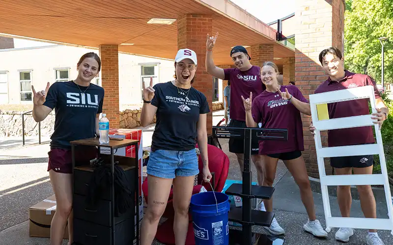 Students posing with move-in supplies, smiling and making hand signs while standing outside a residence hall.