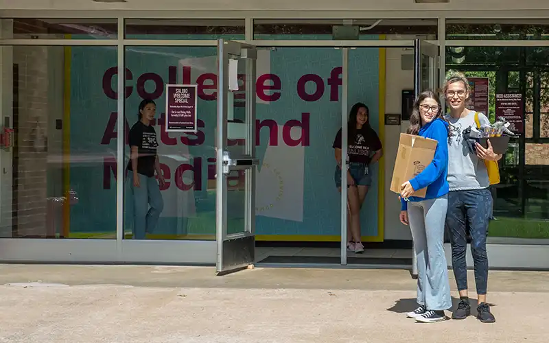 Two students carrying belongings and posing in front of the College of Arts and Media entrance during move-in, with others visible inside the building.