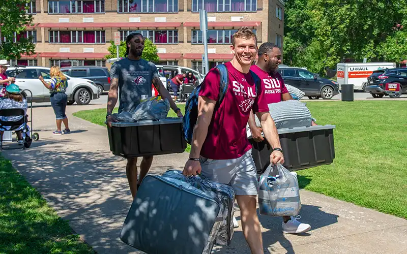 Students carrying bins, bags, and bedding across the lawn during move-in, smiling as they walk toward the residence hall.