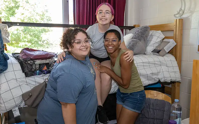 Three students in a residence hall room smiling and posing together beside a lofted bed.